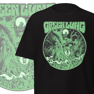 May include: Black t-shirt featuring a green graphic with the text "GREENLUNG" above a circular design. The design includes a tree, figures, a moon, and a lantern. The design is repeated on the left side of the image.
