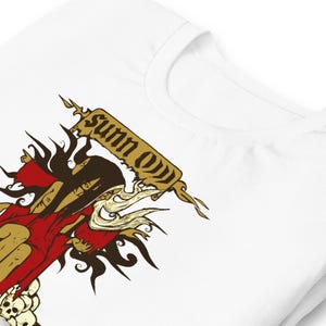 May include: White t-shirt featuring a graphic design with the text "Sunn O))) ". The design includes a figure in red and gold, with skull details. The shirt is made of a soft, comfortable material.