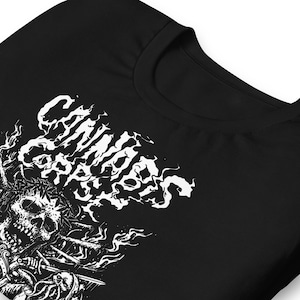 Può includere: T-shirt nero con un design grafico bianco. Il design presenta un teschio, spade e il testo "Cannibal Corpse" in un font stilizzato. La maglietta è realizzata in un materiale morbido.