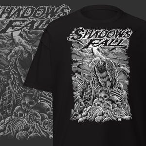 May include: Black t-shirt featuring a detailed grayscale illustration. The design includes a vulture perched atop a pile of skulls and bones, with the words "Shadows Fall" in a stylized font. The artwork has a gothic or metal theme.