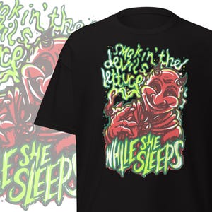 May include: Black t-shirt featuring a red devil cartoon with the text "Smokin' in the devil's lettuce prize" and "While she sleeps" in green and white. The design is a humorous illustration.