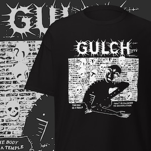 Puede incluir: Camiseta negra con un diseño gráfico blanco. El diseño incluye la palabra "GULCH" sobre una ilustración de pared de ladrillo con calaveras y una figura. El texto en la camiseta dice "THE BODY IS A TEMPLE" y "THAT IS DAMNED TO DEMOLITION."