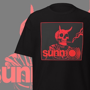 May include: Black t-shirt with a red graphic featuring a skull with horns smoking a cigarette, inside a red square. The word "sunn" is below the skull in red, with a circular design.