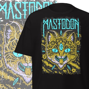 May include: Black t-shirt featuring a yellow and teal graphic of a cat-like creature with three eyes and the band name "MASTODON" in teal. The design has a distressed, cracked effect.