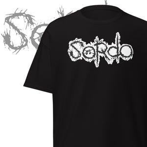 May include: Black t-shirt with the white word "Sordo" in a distressed font. The design includes a musical note within the word. A gray, abstract "S" shape is in the background.