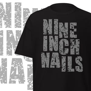 May include: Black t-shirt with the band name "Nine Inch Nails" printed in distressed gray lettering. The text is stacked vertically, with a grunge-style font. The shirt is a classic crew neck.