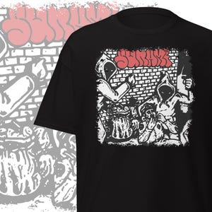 May include: Black t-shirt featuring a graphic design with the word "SURRAX" in red graffiti-style lettering. The design includes hooded figures and a brick wall background, creating a street art aesthetic.