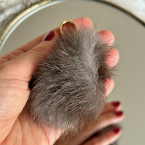 Op de afbeelding: Een pluizige, grijze bontpompon met een goudkleurige metalen ring. De pompon wordt tegen een spiegel gehouden, die de zachte textuur van het bont weerspiegelt. De achtergrond is een zachte, neutrale kleur.