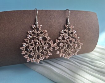 May include: A pair of beige lace earrings with a floral design. The earrings are decorated with small, light pink beads and hang from silver-coloured hooks. The earrings are displayed against a brown background.