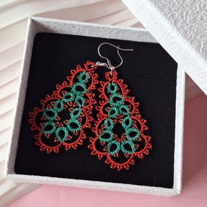 Puede incluir: Un par de pendientes calados hechos a mano en una caja de regalo. Los pendientes presentan un diseño floral rojo y verde. Los pendientes se exhiben en una caja blanca con interior negro, sobre un fondo rosa.