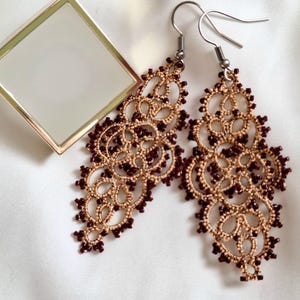 May include: A pair of ornate, beige and burgundy dangle earrings with intricate lace-like detailing. The earrings feature a floral design with small burgundy beads. A gold-framed square mirror is in the background.