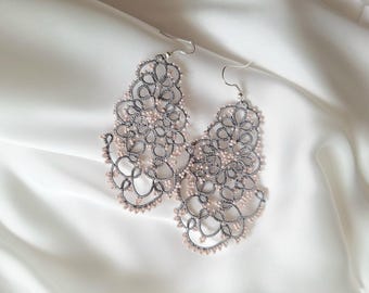 Può includere: Un paio di orecchini traforati color argento con un motivo floreale. Gli orecchini presentano un delicato motivo a pizzo e piccole perline rosa chiaro. Sono presentati su un tessuto bianco e setoso.
