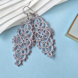May include: A pair of silver-colored, openwork lace earrings with a floral design. The earrings are displayed on a light blue surface with a white fabric and a small, square box with a gold trim.
