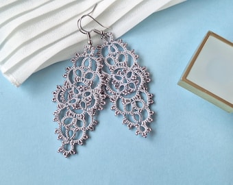 Può includere: Un paio di orecchini in pizzo traforato color argento con un motivo floreale. Gli orecchini sono esposti su una superficie azzurra con un tessuto bianco e una piccola scatola quadrata con bordi dorati.