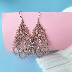 Può includere: Un paio di orecchini delicati color oro con un design intricato e traforato. Gli orecchini sono appesi a ganci argentati.