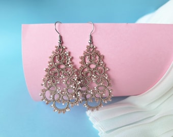 May include: A pair of delicate, gold-toned earrings with an intricate, lacy design. The earrings are hanging from silver hooks.
