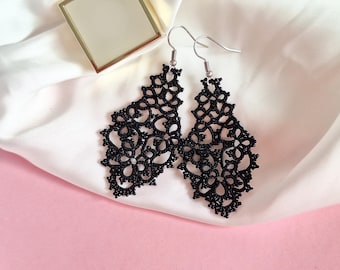 May include: A pair of black, openwork drop earrings with a floral pattern. The earrings are crafted from a delicate, lace-like material and feature silver-coloured hooks. They are presented on white satin fabric against a pink backdrop.