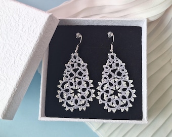 May include: Delicate, openwork teardrop earrings in a white gift box with a black interior. The earrings are a light, silvery colour with intricate detailing and small white accents. The box is open, revealing the earrings, with a soft, white fabric in the background.