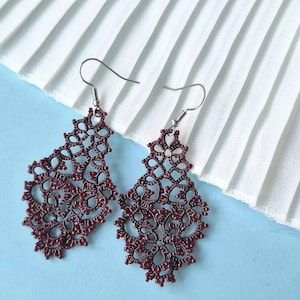 May include: A pair of burgundy-colored, open-work earrings with a floral design. The earrings are made of a delicate, lace-like material and hang from silver-colored hooks. The background is a light blue surface with a white pleated fabric.