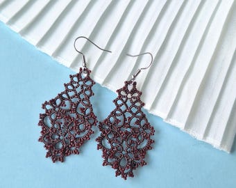 May include: Two burgundy-coloured, openwork earrings with a floral design. The earrings are made of a delicate, lace-like material and hang from silver-coloured hooks. The background is a light blue surface with white pleated fabric.