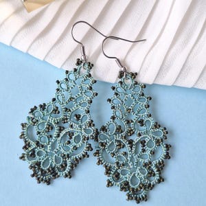 Op de afbeelding: Een paar sierlijke, lichtblauwe kanten oorbellen met donkerbruine kraalaccenten. De oorbellen hebben een bloemenpatroon en zijn opgehangen aan zilverkleurige oorhaken. De achtergrond is een lichtblauwe ondergrond met witte, geplooide stof.