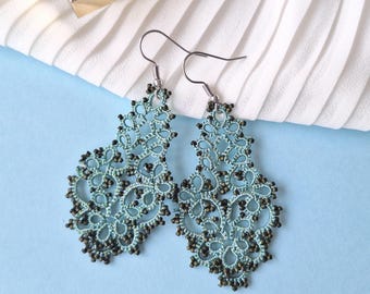 May include: A pair of delicate, pale blue lace earrings with dark brown bead detailing. The earrings feature a floral pattern and are hung from silver-tone ear wires. The background is a light blue surface with white, pleated fabric.