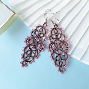 May include: A pair of dangle earrings with a dark brown filigree design and small pink beads. The earrings are set against a light blue background with a white pleated fabric and a small gold box.