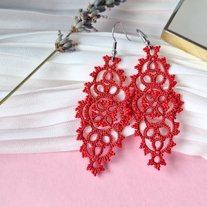 May include: A pair of vibrant red, intricately designed dangle earrings. The earrings feature a delicate, lace-like pattern with a floral motif. They hang from silver-colored hooks. The background includes a sprig of lavender and a small gold-trimmed box.