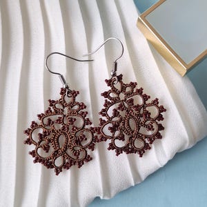 May include: A pair of brown, intricately designed earrings with a floral pattern. The earrings are made of a delicate material and hang from silver-coloured hooks. They rest on a white, pleated fabric.