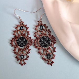 May include: A pair of dangle earrings featuring a detailed, openwork design. The earrings are primarily a reddish-brown color with black accents in the center. The earrings have a floral-like pattern and are suspended from silver-colored hooks.