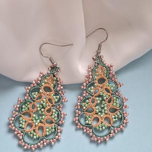 Op de afbeelding: Een paar handgemaakte oorhangers. De oorbellen hebben een gedetailleerd, open ontwerp in tinten van teal, perzik en roze. Het ingewikkelde patroon is geaccentueerd met kleine kralen en hangt aan zilverkleurige haken.