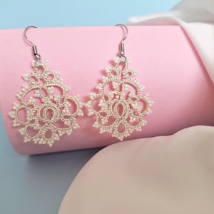 Peut inclure: Une paire de boucles d'oreilles en dentelle crème, au design complexe. Les boucles d'oreilles sont suspendues à des crochets argentés et sont présentées sur un fond rose avec un arrière-plan bleu. Le motif de la dentelle présente des motifs floraux et géométriques.