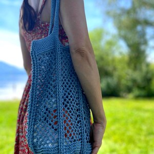 May include: A blue crocheted tote bag with a net-like design. The bag has a rounded bottom and two handles. The bag is being held outdoors on a sunny day.