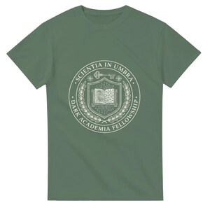 Könnte beinhalten: Ein salbeigrünes T-Shirt mit kurzen Ärmeln und einem weißen kreisförmigen Emblem auf der Brust. Das Emblem zeigt ein offenes Buch, einen Schlüssel und Lorbeerzweige mit dem Text "SCIENTIA IN UMBRA" und "DARK ACADEMIA FELLOWSHIP" im Rand.