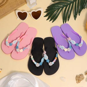 Puede incluir: Una colección de chanclas coloridas en rosa, negro y morado, adornadas con detalles decorativos de conchas marinas y cuentas. Las sandalias están dispuestas con gafas de sol, un sombrero de paja y follaje tropical, sugiriendo un tema de playa.