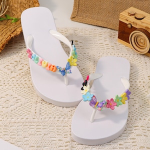 Puede incluir: Chanclas blancas con adornos florales y mariposas de colores. Las sandalias tienen una correa blanca y una suela acolchada. Las flores y mariposas son en tonos rosa, amarillo, azul y verde.