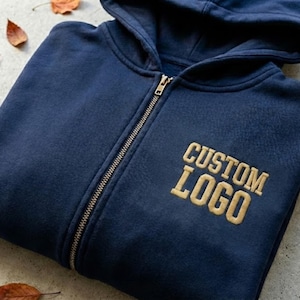May include: A navy blue zip-up hoodie with a hood and a gold-colored "CUSTOM LOGO" embroidered on the chest. The zipper is silver. The hoodie is lying flat on a surface with a few fallen leaves.