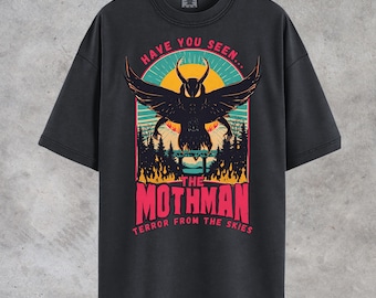 May include: Dark gray t-shirt featuring a graphic of the Mothman with the text "HAVE YOU SEEN... THE MOTHMAN TERROR FROM THE SKIES". The design includes a black silhouette of the Mothman, a sunset, and a forest.