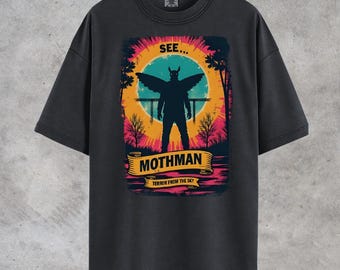 May include: Dark gray t-shirt featuring a colorful graphic of the Mothman, a cryptid with wings and horns, set against a sunset backdrop. The text "SEE... MOTHMAN" is displayed above the figure, with "TERROR FROM THE SKY" below.