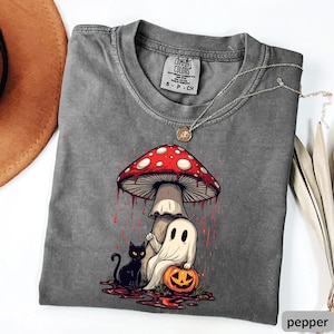 May include: A gray Comfort Colors t-shirt featuring a whimsical Halloween design. The graphic includes a red and white mushroom, a ghost, a black cat, and a carved pumpkin. A silver necklace with a pendant is draped across the shirt.