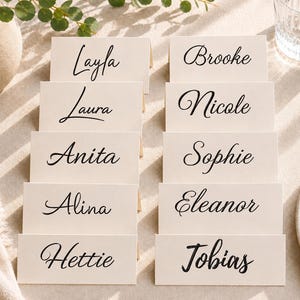 May include: Ten off-white rectangular folded place cards arranged in two columns. Each card features a unique name printed in a black cursive script font. The names include Layla, Brooke, Laura, Nicole, Anita, Sophie, Alina, Eleanor, Hettie, and Tobias.