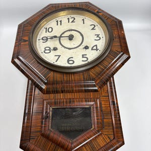 May include: Vintage wall clock with a patterned wooden case in brown and black. The clock face is white with black numerals and hands. The manufacturer's name, "Seikosha," is displayed on the lower section of the clock.