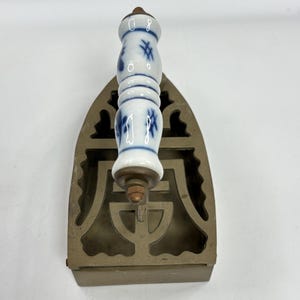 May include: Antique metal flat iron with a decorative handle. The handle is white porcelain with blue floral designs. The iron has a rectangular base with an openwork design.