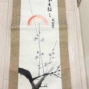 May include: A traditional Japanese scroll painting featuring calligraphy and a branch with blossoms. The artwork includes a red sun and black ink brushstrokes. The scroll has a gold patterned border and is displayed vertically.
