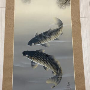 May include: Japanese scroll painting featuring two koi fish swimming in a gray-toned body of water. The artwork includes a detailed depiction of pine branches at the top. The scroll has a decorative border.