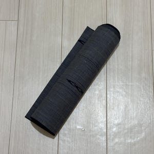 May include: A charcoal gray roll of fabric featuring a subtle, fine-lined horizontal texture. The material is tightly coiled into a cylindrical shape, displaying a consistent dark gray tone throughout the visible surface of the textile roll.