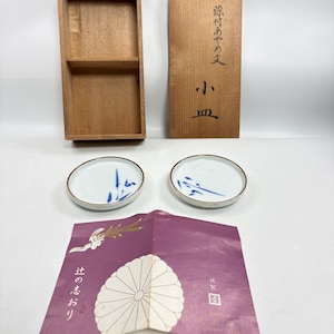 Può includere: Due piccoli piatti rotondi in ceramica bianca con motivi di bambù dipinti in blu, una scatola rettangolare in legno con un coperchio recante calligrafia giapponese e un inserto in carta viola piegato con un emblema floreale bianco e testo giapponese.