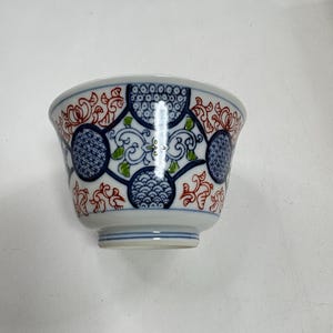 May include: A ceramic bowl featuring a white base with a repeating pattern of blue circular medallions containing wave and dot motifs, interspersed with red and green floral scrollwork designs. The bowl has a flared rim and a small footed base.