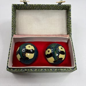 May include: Two round, dark blue meditation balls featuring hand-painted panda bears and green bamboo stalks, presented in a rectangular, fabric-covered box with a green floral pattern and a bright red interior lining for storage.