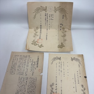 以下が含まれることがあります： オフホワイトの紙に黒い印刷文字、上部に鳳凰のイラスト、下部に竹のモチーフが描かれた3枚の古い日本の書類。各書類には赤い四角い印鑑が押されており、ページ全体に正式な日本語の文字が記載されています。